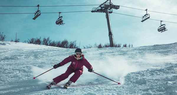 En person i full fart ned en alpinbakke. I bakgrunnen ser man en stolheis. Alpinisten er en kvinne og har på seg hjelm.