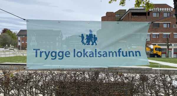 Et skilt der det står "Trygge lokalsamfunn" er hengt opp mellom en lyktestolpe og et tre på torget foran Rådhuset i Ski. Foto.