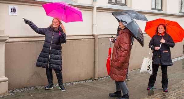 Tre damer med hver sin paraply står og peker mot et skilt på en husvegg.