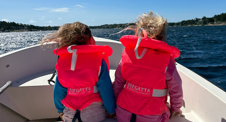 To jenter med redningsvest sitter i en båt og ser fremover. Vi ser ryggene deres.